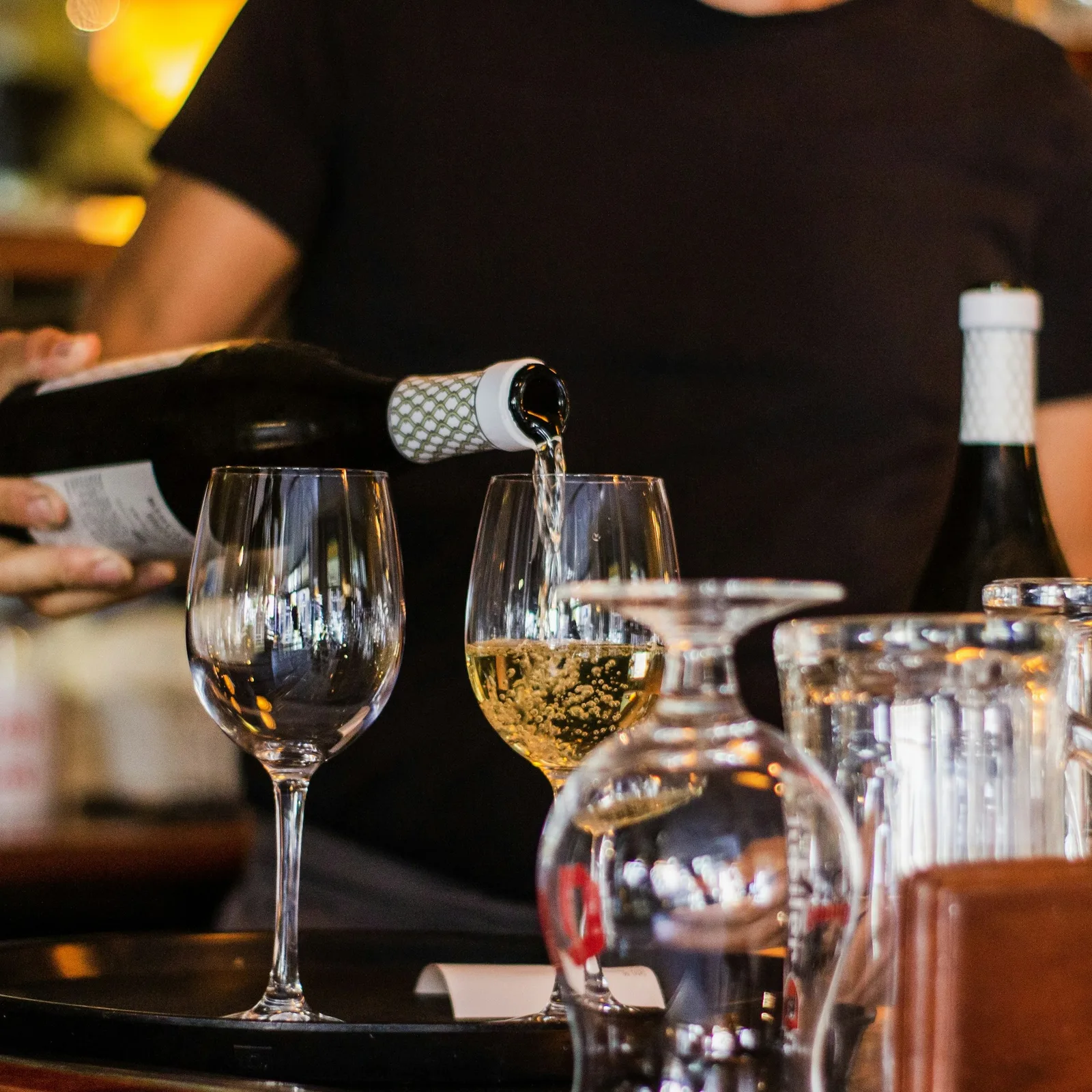 A person pouring a bottle of sparkling wine into a glass at a bar or restaurant, with several other glasses and bottles on the counter